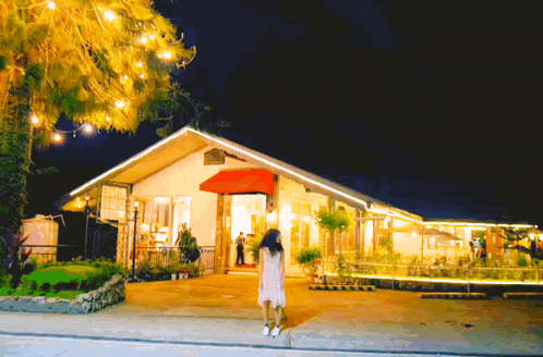 A Woman In A White Dress Is Standing In Front Of A House With A Red Awning GIF