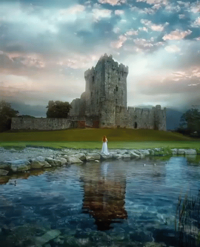 A Woman In A White Dress Is Standing In Front Of A Large Stone Castle GIF