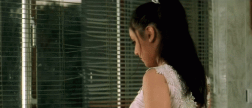 A Woman In A White Dress Is Standing In Front Of A Window With Blinds On It . GIF