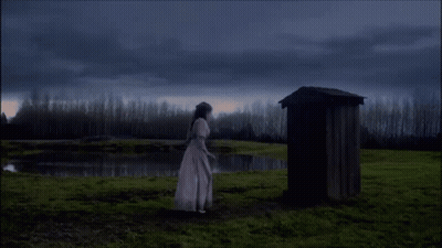 A Woman In A White Dress Is Standing In Front Of A Wooden Outhouse GIF