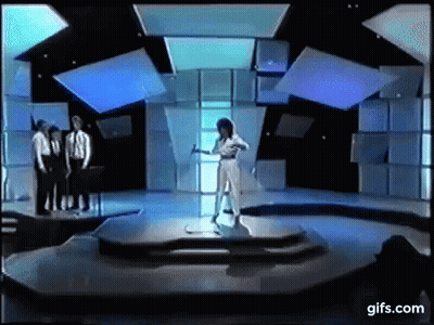 A Woman In A White Dress Is Standing On A Stage In Front Of A Group Of Men In Suits . GIF