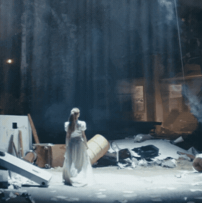 A Woman In A White Dress Is Standing On A Stage With A Barrel In The Background GIF