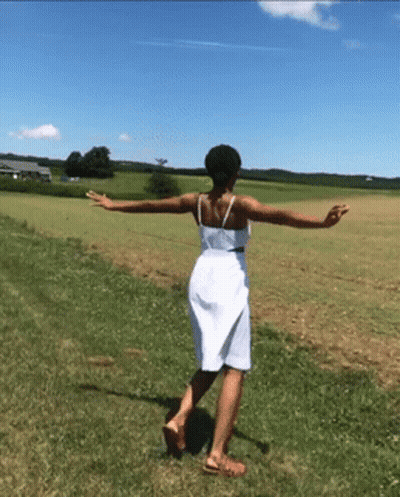 A Woman In A White Dress Is Walking Through A Grassy Field GIF