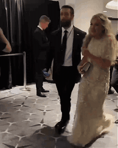 A Woman In A White Dress Is Walking With A Man In A Black Suit GIF