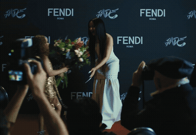 A Woman In A White Dress Stands In Front Of A Wall That Says Fendi Roma GIF