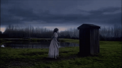 A Woman In A White Dress Stands In Front Of A Wooden Outhouse In A Field GIF