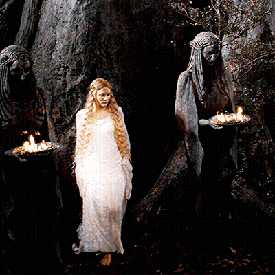 A Woman In A White Dress Stands In Front Of Two Statues Holding Candles GIF