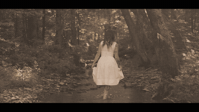 A Woman In A White Dress Walks Through The Woods GIF