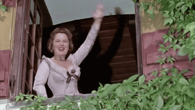 A Woman In A White Dress Waving From A Balcony GIF