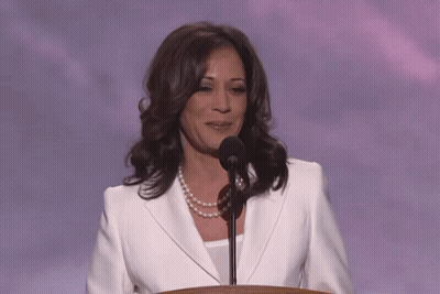 A Woman In A White Jacket And Pearl Necklace Stands At A Podium Speaking Into A Microphone GIF