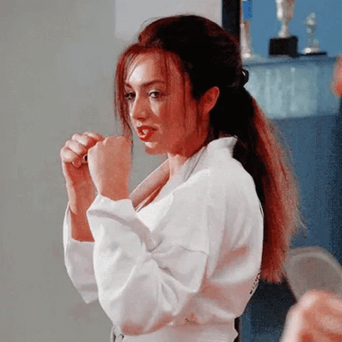 A Woman In A White Karate Uniform Is Standing In A Room With Her Hands In Her Fist . GIF