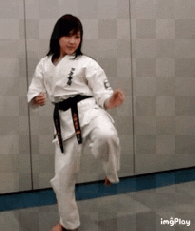 A Woman In A White Karate Uniform With A Black Belt Is Kicking A Ball . GIF