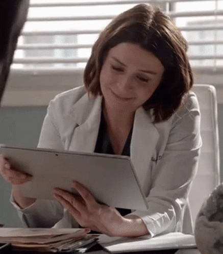 A Woman In A White Lab Coat Is Sitting At A Desk Holding A Tablet Computer . GIF