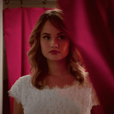 A Woman In A White Lace Top Is Looking Through A Pink Curtain GIF