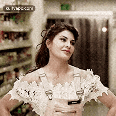 A Woman In A White Off The Shoulder Top And Pink Overalls Is Standing With Her Hands On Her Hips In A Store . GIF