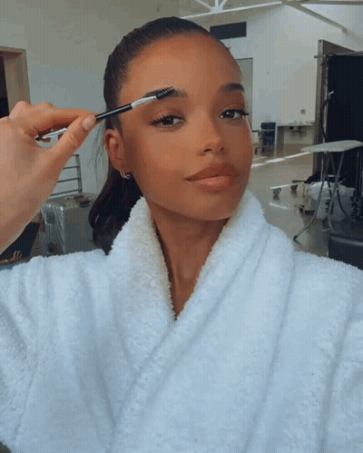 A Woman In A White Robe Has Her Eyebrows Brushed By Someone GIF