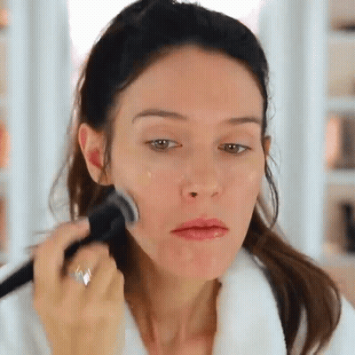 A Woman In A White Robe Is Applying Makeup To Her Face GIF