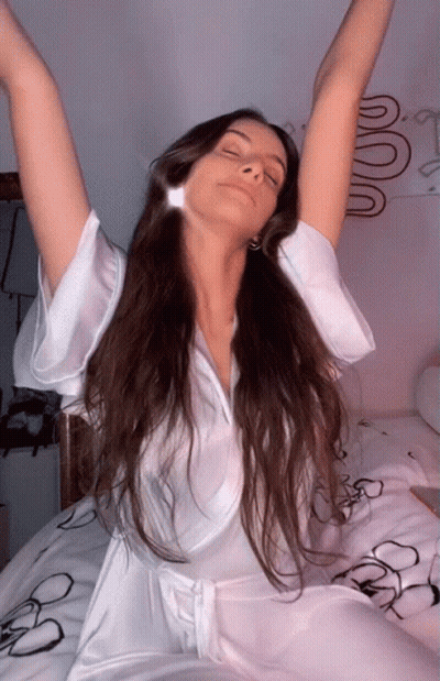 A Woman In A White Robe Is Sitting On A Bed With Her Eyes Closed GIF