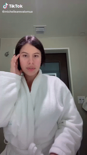 A Woman In A White Robe Is Standing In Front Of A Mirror With A Tiktok Logo Above Her GIF
