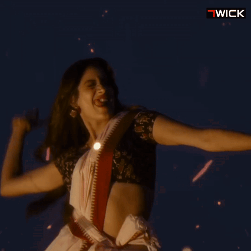 A Woman In A White Saree Is Dancing In Front Of A Sign That Says " Wick " GIF
