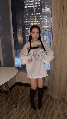 A Woman In A White Shirt And Black Boots Stands In Front Of A Window With A City In The Background GIF