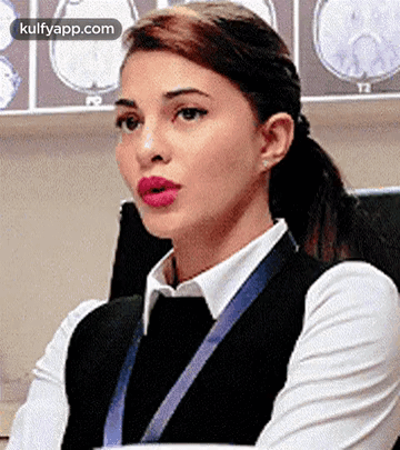 A Woman In A White Shirt And Black Vest Is Sitting In Front Of A Wall With Brain Scans . GIF