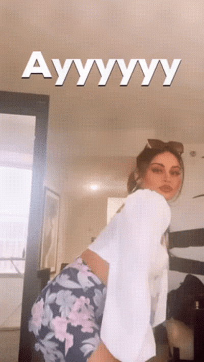 A Woman In A White Shirt And Floral Shorts Is Standing In A Room With The Words Ayyyyyy Written On The Ceiling Above Her GIF