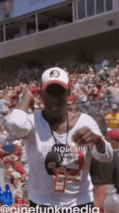 A Woman In A White Shirt And Hat Is Standing In Front Of A Crowd Of People . GIF