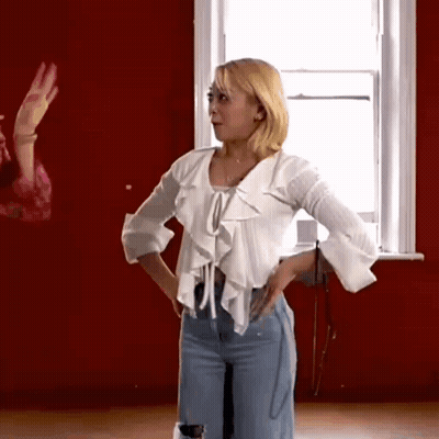 A Woman In A White Shirt And Jeans Is Standing In Front Of A Window . GIF