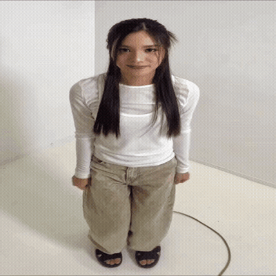 A Woman In A White Shirt And Khaki Pants Is Standing In A White Room GIF