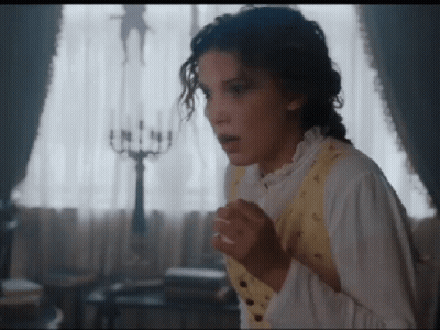 A Woman In A White Shirt And Yellow Vest Is Standing In Front Of A Window With Her Hands On Her Chest . GIF