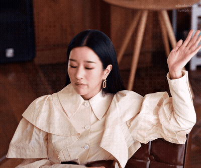 A Woman In A White Shirt Is Sitting In A Chair With Her Hand Up In The Air GIF