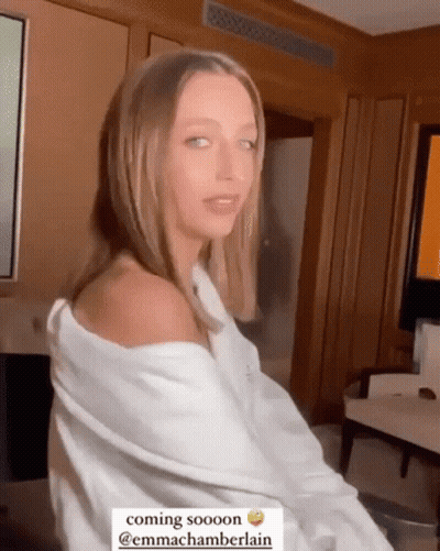 A Woman In A White Shirt Is Sitting In A Room With A Caption That Says Coming Sooooon @Emmachamberlain GIF