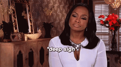 A Woman In A White Shirt Is Sitting In Front Of A Dresser And Flowers And Says Deep Sigh GIF