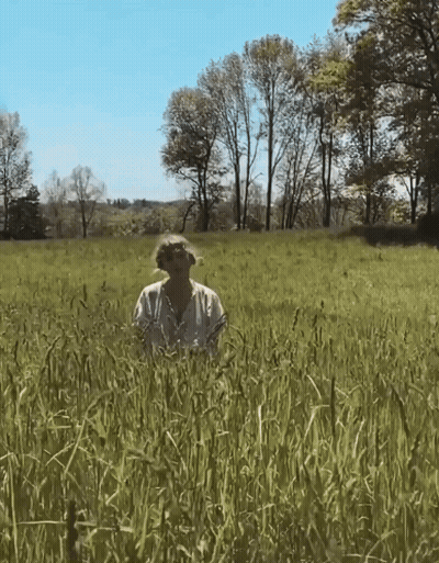 A Woman In A White Shirt Is Standing In A Grassy Field GIF