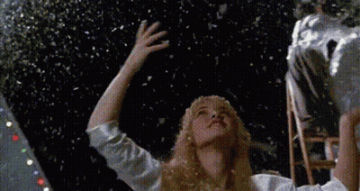 A Woman In A White Shirt Is Standing In Front Of A Christmas Tree With Snow Falling From The Sky GIF