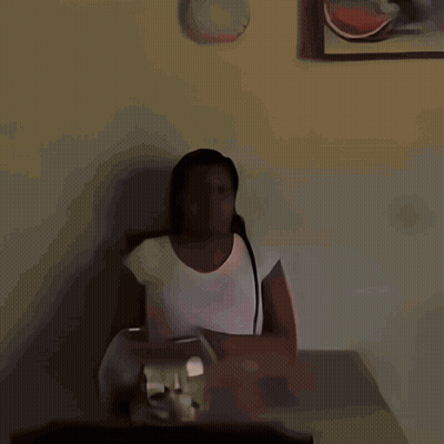 A Woman In A White Shirt Sits At A Table With A Clock On The Wall Above Her GIF