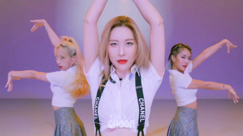 A Woman In A White Shirt With Chanel Suspenders Is Standing In Front Of Two Other Women . GIF