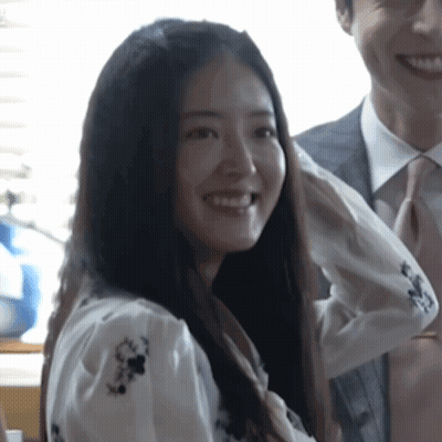 A Woman In A White Shirt With Flowers On It Smiles Next To A Man In A Suit And Tie GIF