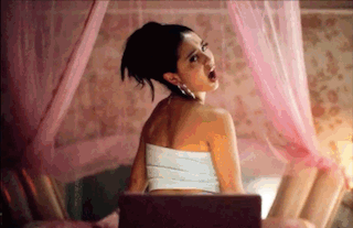 A Woman In A White Strapless Top Is Sitting On A Bed With A Pink Canopy . GIF
