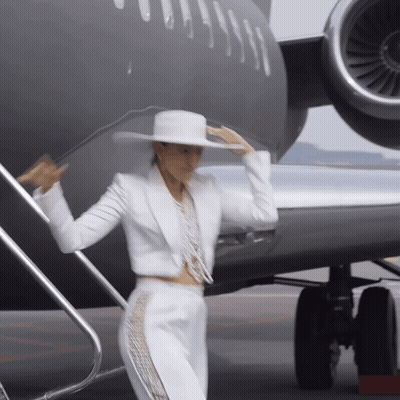 A Woman In A White Suit And Hat Is Standing In Front Of A Plane GIF