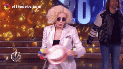 A Woman In A White Suit Is Holding A Tambourine In Front Of A Screen That Says Eltrecetv.com GIF