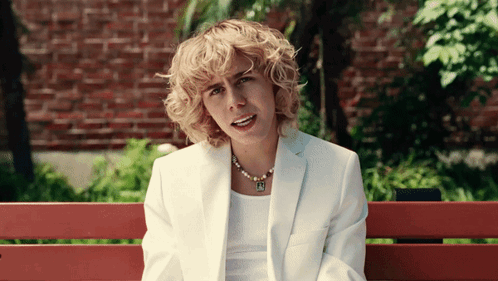 A Woman In A White Suit Is Sitting On A Bench With A Brick Wall In The Background GIF