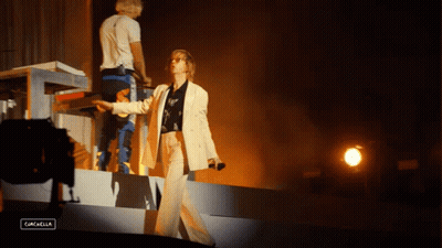 A Woman In A White Suit Stands On A Stage In Front Of A Sign That Says Gascella GIF