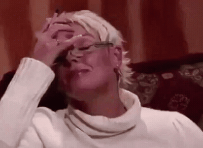 A Woman In A White Sweater And Glasses Is Sitting On A Couch Covering Her Face With Her Hand . GIF