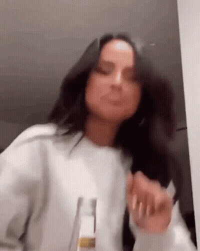 A Woman In A White Sweater Is Dancing In A Room With A Bottle In Her Hand . GIF