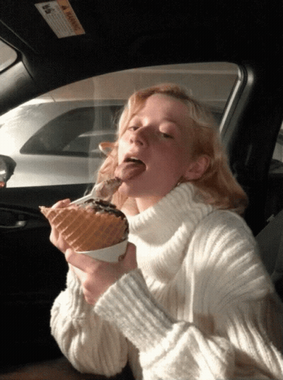 A Woman In A White Sweater Is Eating An Ice Cream Cone In A Car GIF