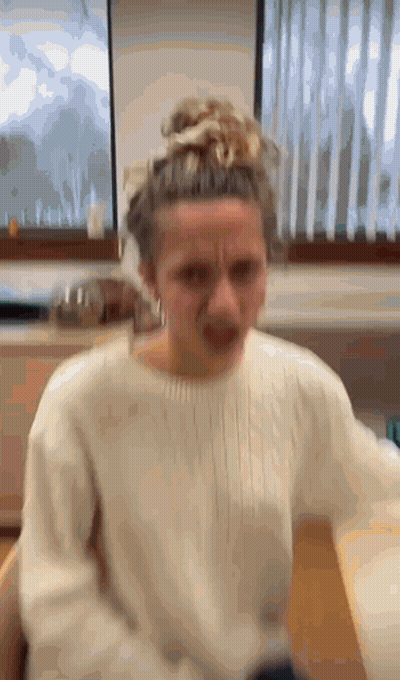 A Woman In A White Sweater Is Making A Funny Face In Front Of A Window . GIF
