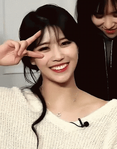 A Woman In A White Sweater Is Smiling And Giving A Peace Sign GIF
