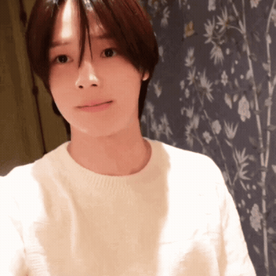 A Woman In A White Sweater Stands In Front Of A Wall With Flowers On It GIF